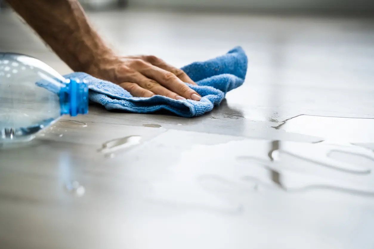 Hand cleaning a water spill off the floor with a blue towel. Find out how to reduce excess moisture and humidity in your home to prevent mold growth.