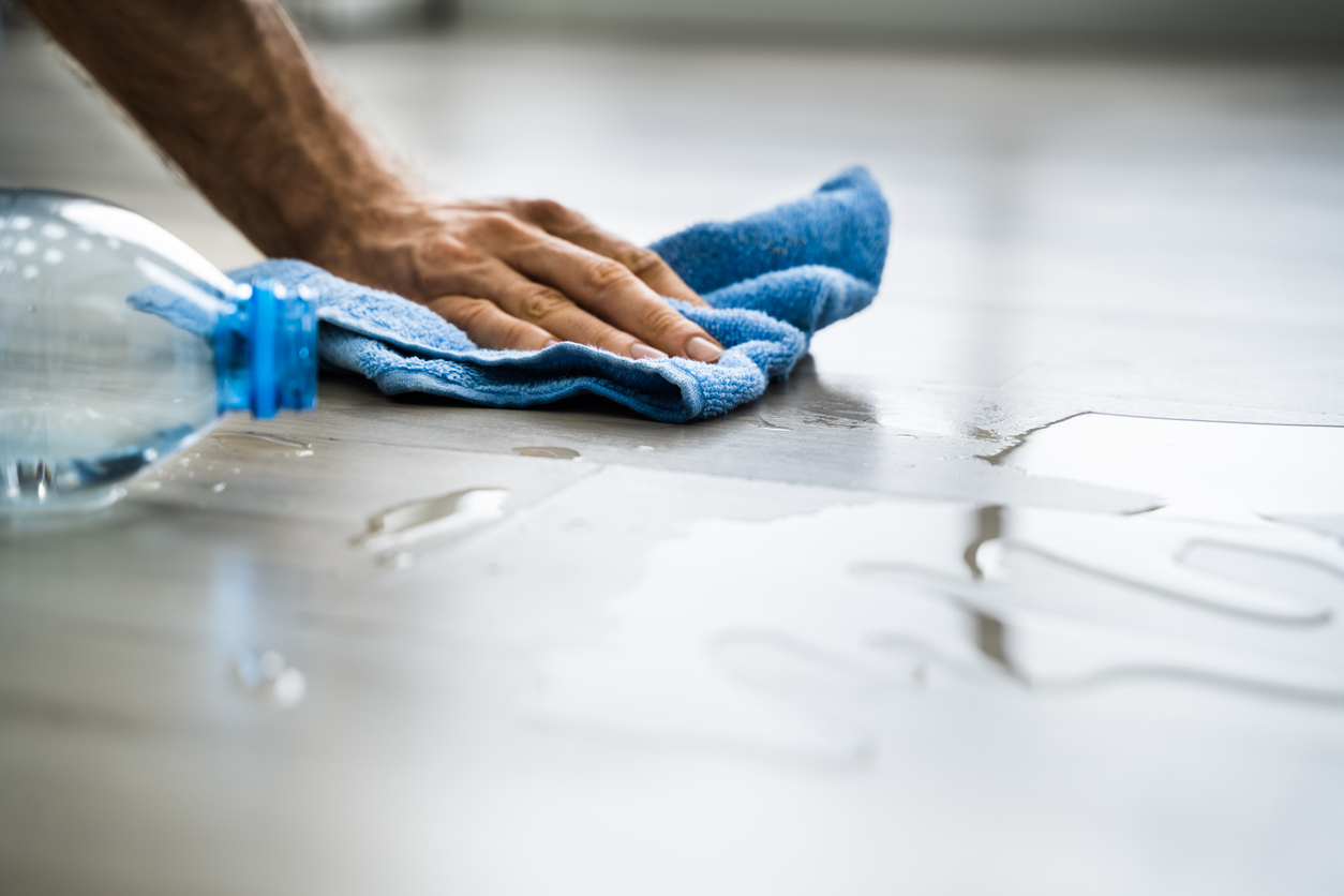 Hand cleaning a water spill off the floor with a blue towel. Find out how to reduce excess moisture and humidity in your home to prevent mold growth.